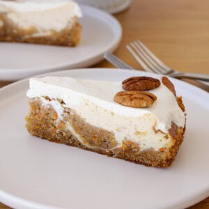 A slice of carrot cake cheesecake with a light brown base, creamy white frosting, and two whole pecans on top, served on a white plate. A fork and another plate with cake are in the background.