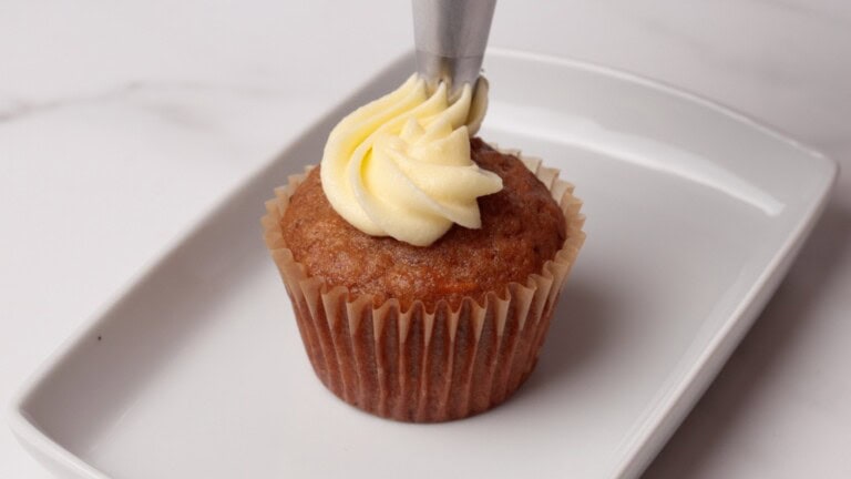 A cupcake in a brown paper liner is being decorated with creamy cream cheese frosting using a piping bag on a rectangular white plate.