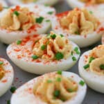 Close-up of deviled eggs topped with paprika and chopped chives, arranged on a gray surface. The creamy yolk filling is piped into the egg whites and garnished for a colorful, appetizing appearance.