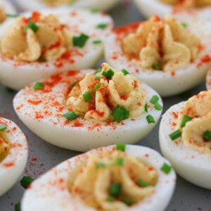 Close-up of deviled eggs topped with paprika and chopped chives, arranged on a gray surface. The creamy yolk filling is piped into the egg whites and garnished for a colorful, appetizing appearance.