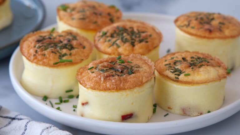 Six fluffy egg bites topped with chopped herbs are arranged on a white plate. The egg bites have a golden-brown top and creamy yellow sides, with small pieces of vegetables visible inside.