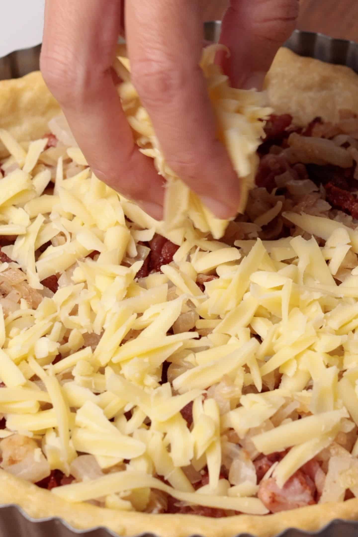 A hand sprinkles shredded cheese over a pie crust filled with chopped ingredients, preparing it for baking.