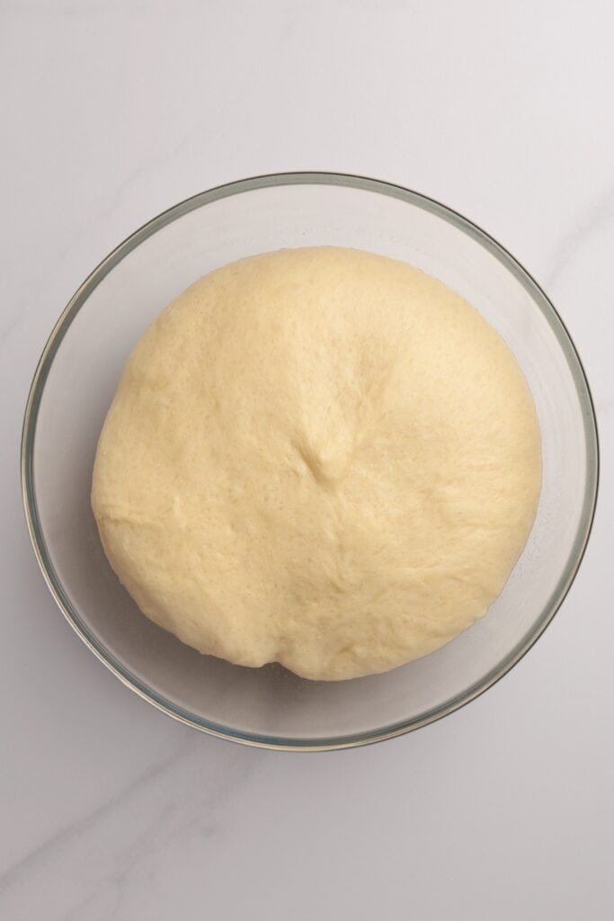 A glass bowl filled with smooth, risen bread dough sits on a light marble surface.