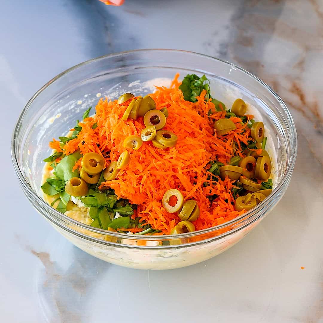 A glass bowl filled with a muffin mix-ins including shredded carrots, sliced green olives, and chopped leafy greens on a marble surface.