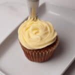 A cupcake in a paper liner is being decorated with a swirl of white frosting using a piping bag on a white rectangular plate.