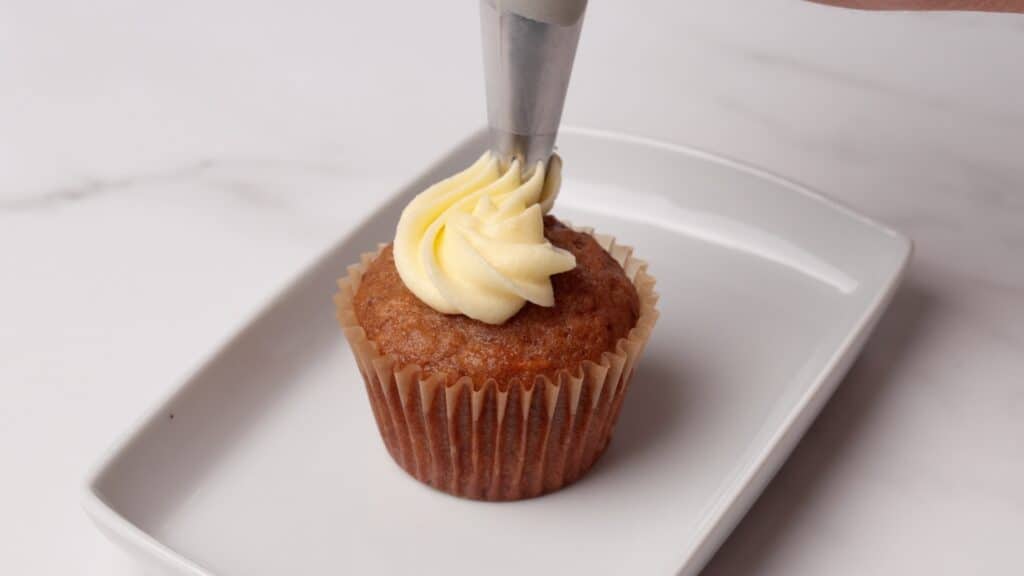 A hand pipes creamy white frosting onto a cupcake with a piping bag, on a rectangular white plate set on a white marble surface.