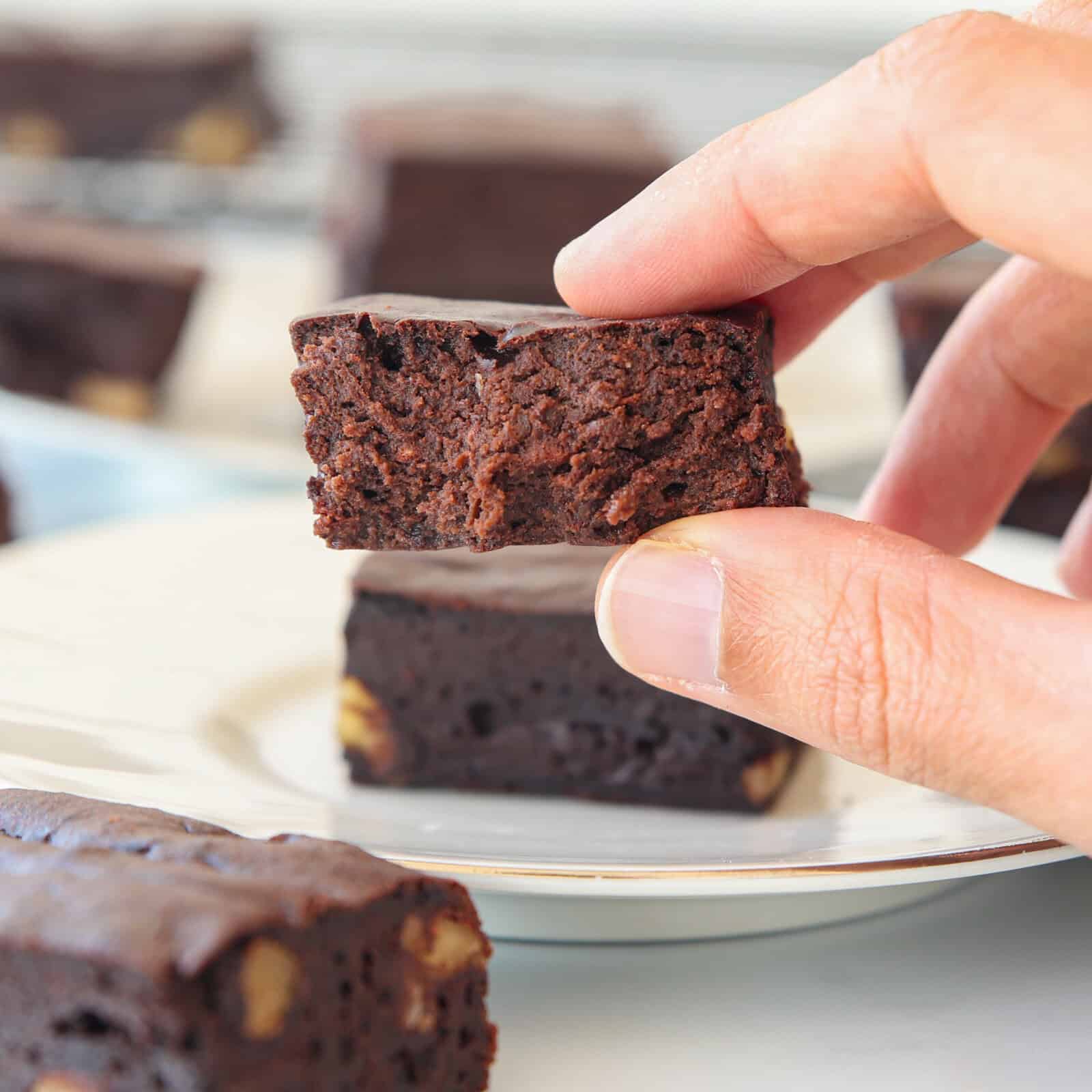 A hand holding a square piece of chocolate brownie above a plate with more brownies. The brownies are dense and fudgy, with a few pieces of nuts visible.