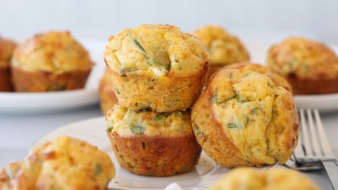 Three savory muffins with visible green herbs are stacked on a white plate, with more muffins in the background and a fork to the right. The muffins appear golden brown and freshly baked.