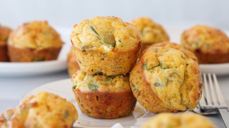 Three savory muffins with visible green herbs are stacked on a white plate, with more muffins in the background and a fork to the right. The muffins appear golden brown and freshly baked.