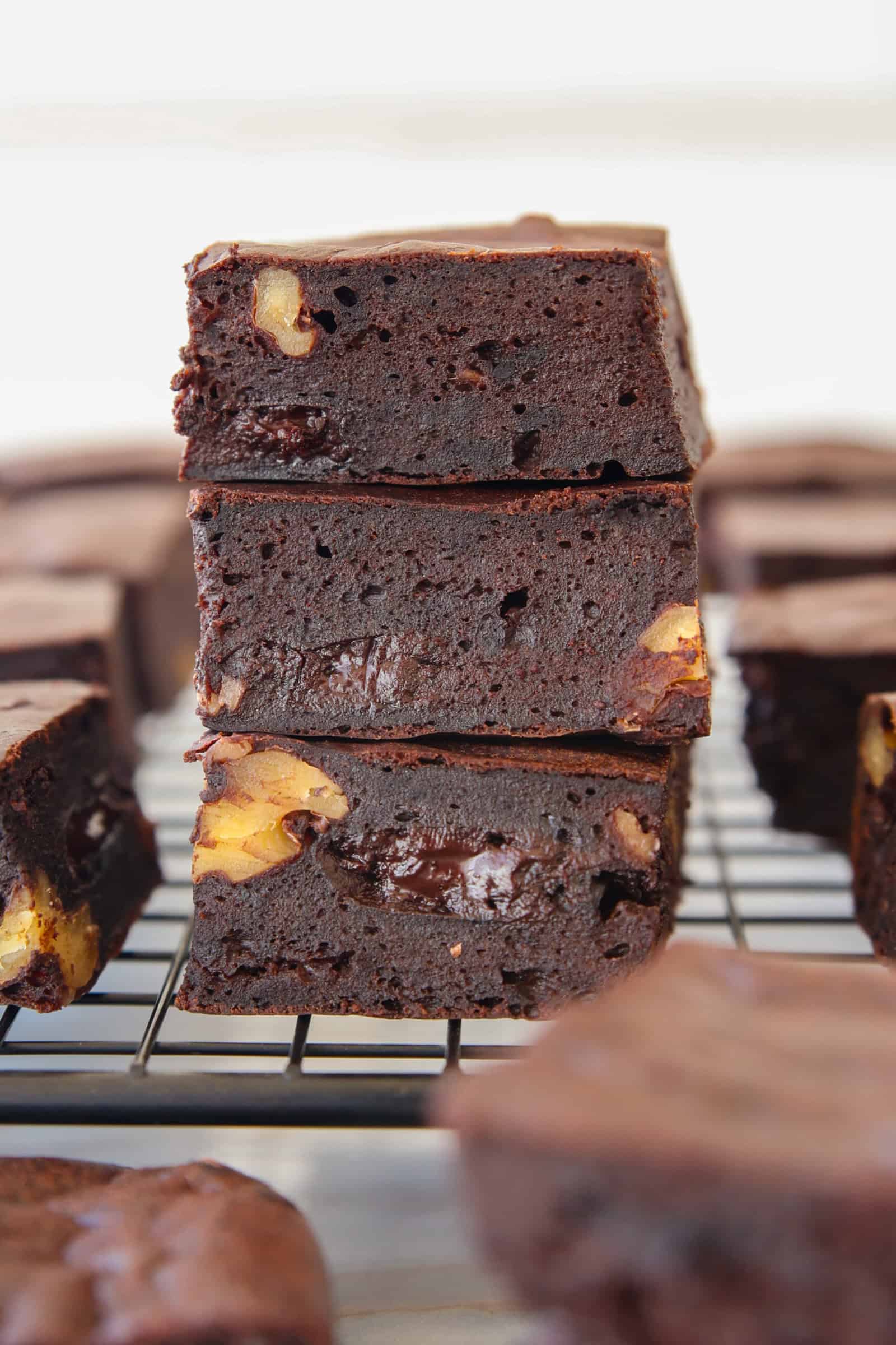 Three thick, fudgy cottage cheese chocolate brownies with visible walnut pieces are stacked on top of each other on a cooling rack, surrounded by more brownies in the background.
