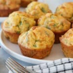 A plate of savory muffins with golden tops and green herb flecks, placed beside a checkered cloth napkin and two forks on a light surface. More muffins are visible in the background.