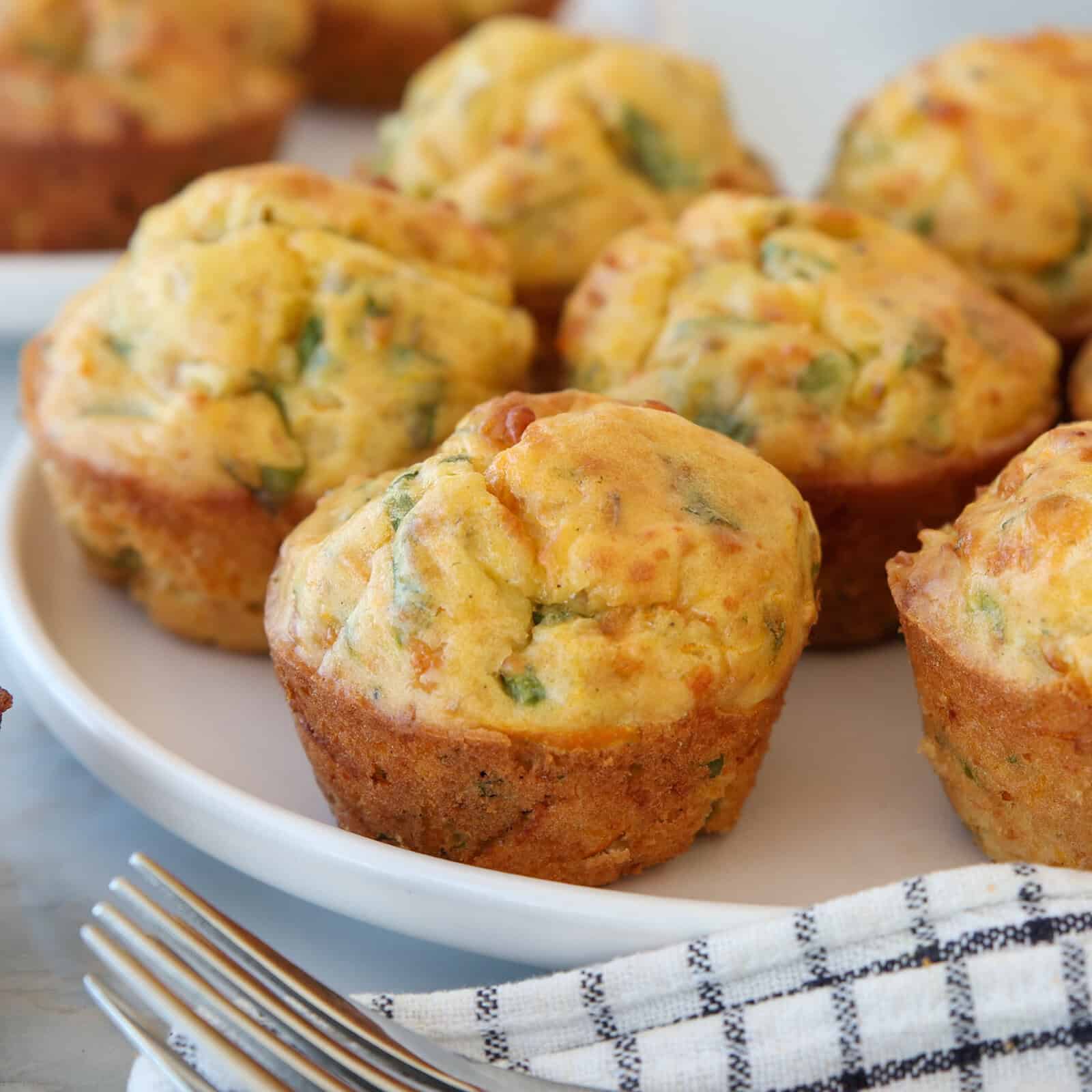 A plate of savory muffins with golden tops and green herb flecks, placed beside a checkered cloth napkin and two forks on a light surface. More muffins are visible in the background.