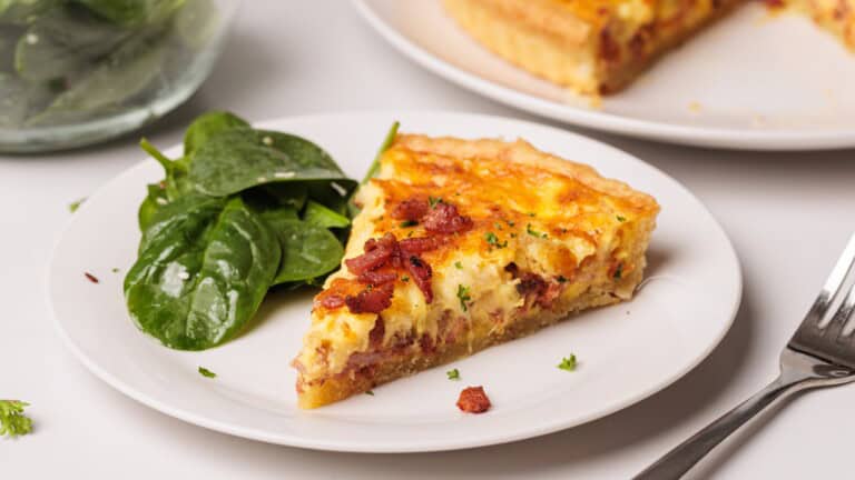 A slice of quiche Lorraine topped with bacon bits and herbs is served on a white plate next to fresh spinach leaves; a fork lies nearby and the rest of the quiche is visible in the background.