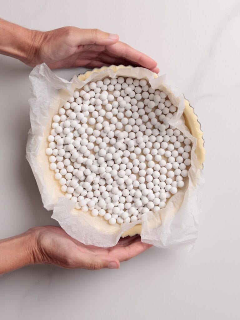 Two hands hold a pie crust lined with parchment paper and filled with white ceramic baking beans, ready for blind baking on a light surface.
