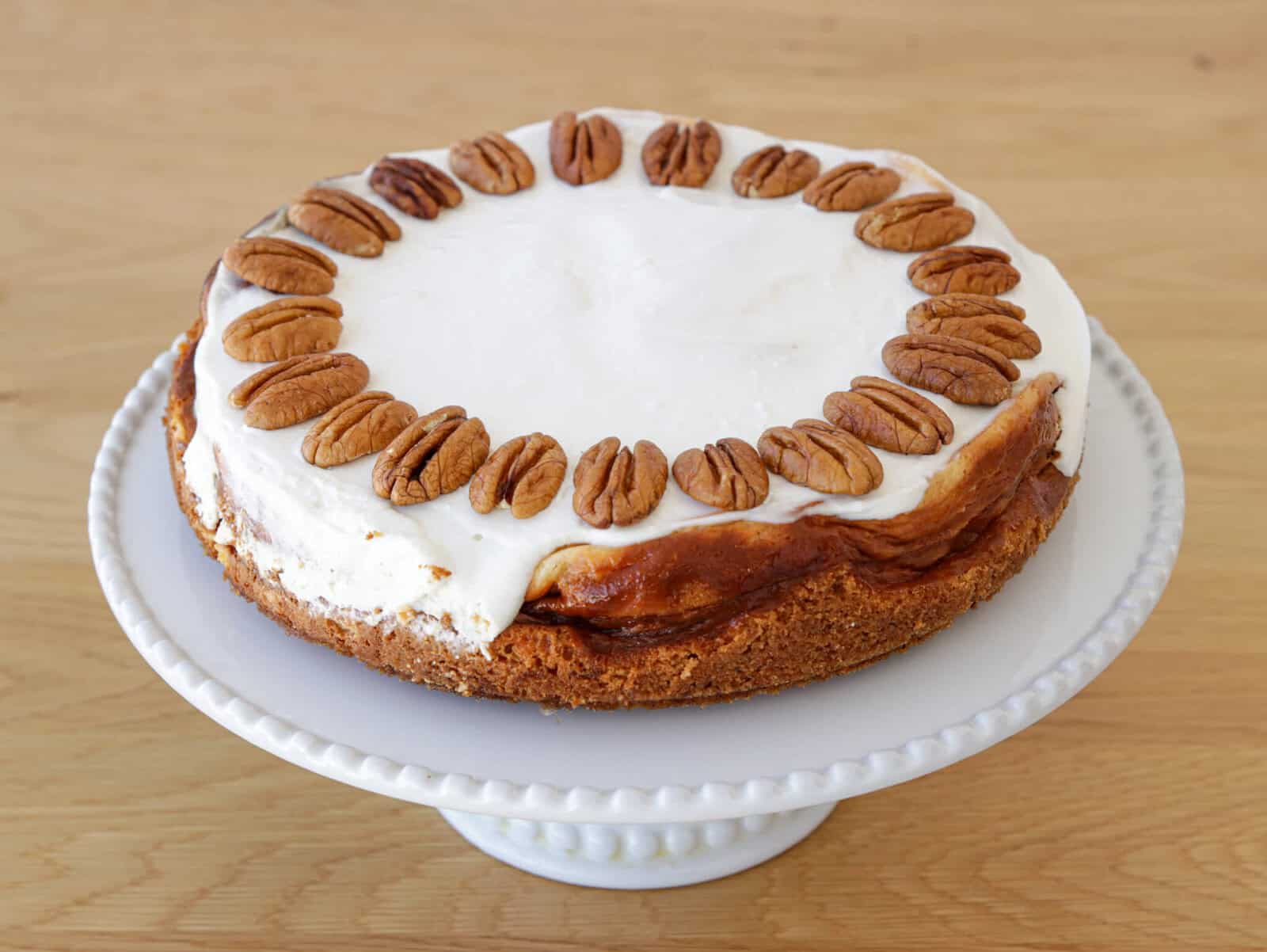 A cheesecake with a golden brown crust, topped with a thick layer of white frosting and decorated with a ring of whole pecans, sits on a white pedestal cake stand on a wooden surface.