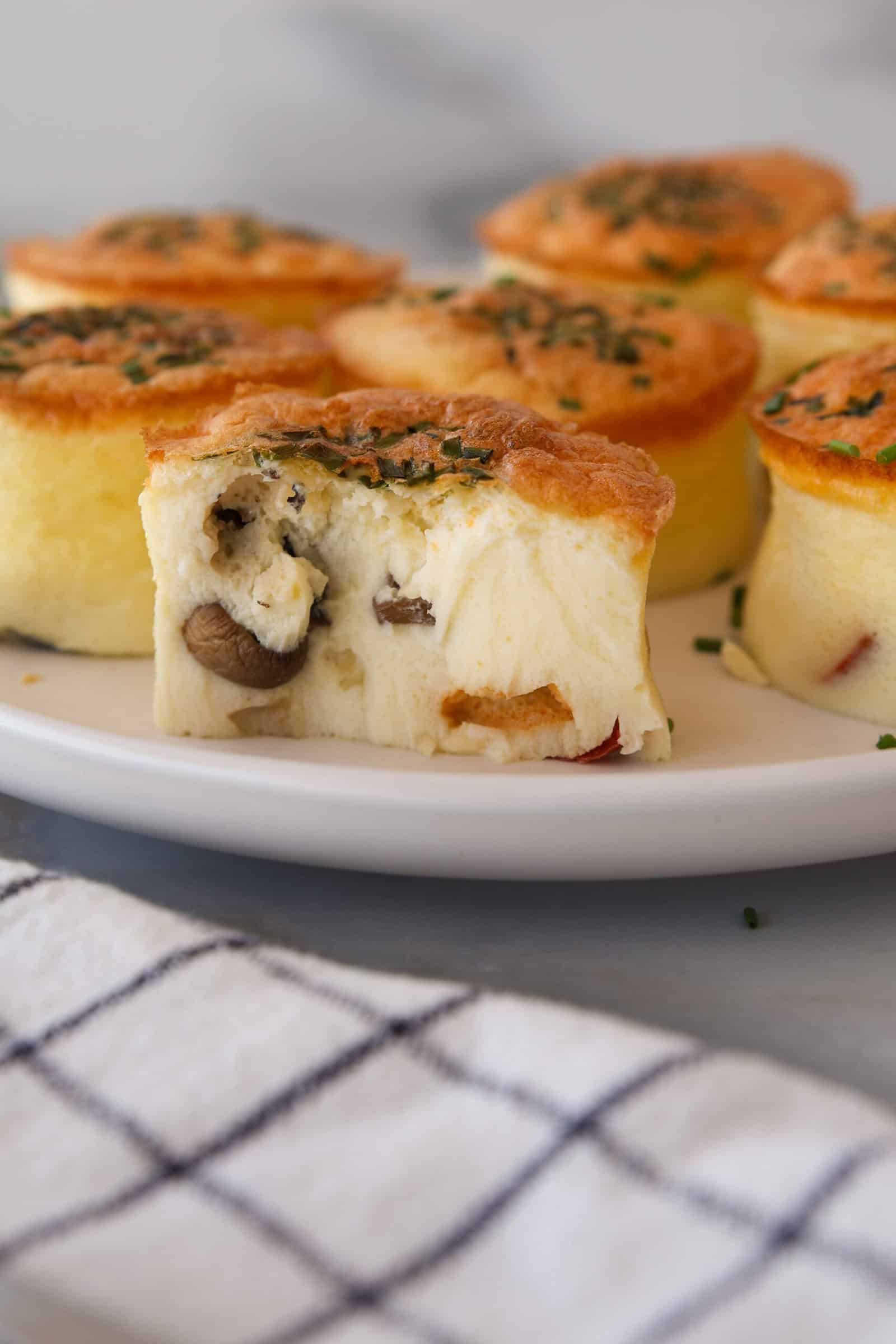 A plate of fluffy, savory cottage cheese egg bite with mushrooms and herbs, one with a bite taken out showing the soft interior. A checkered cloth is in the foreground.