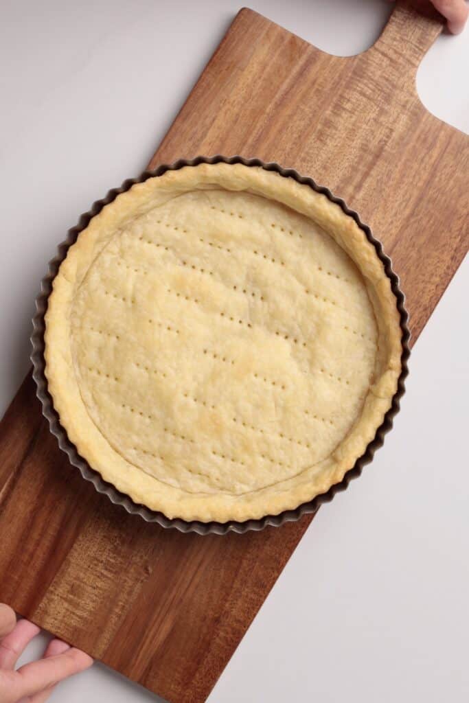 A baked tart crust in a round fluted pan sits on a wooden cutting board, held at one end by a hand. The crust is golden and evenly pricked with fork marks.