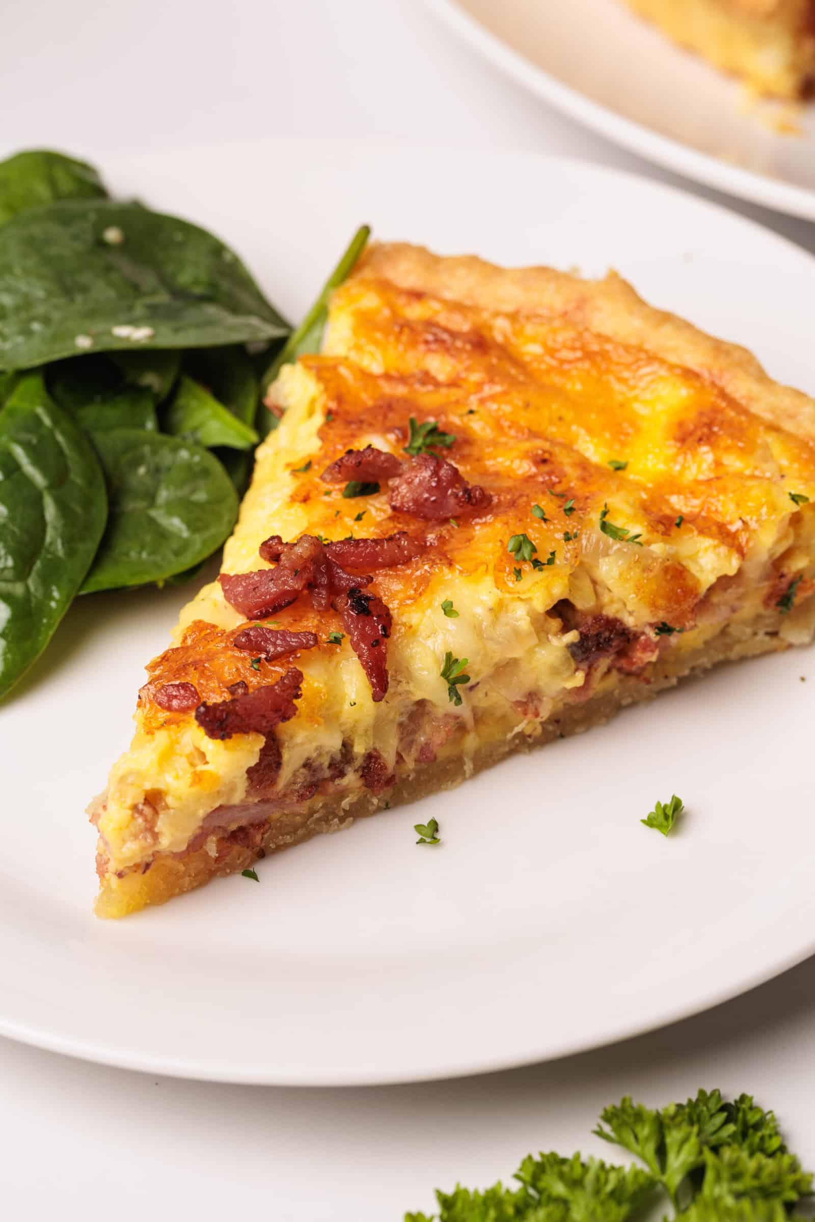 A slice of quiche Lorraine topped with bacon pieces and herbs is served on a white plate with a side of fresh spinach leaves.
