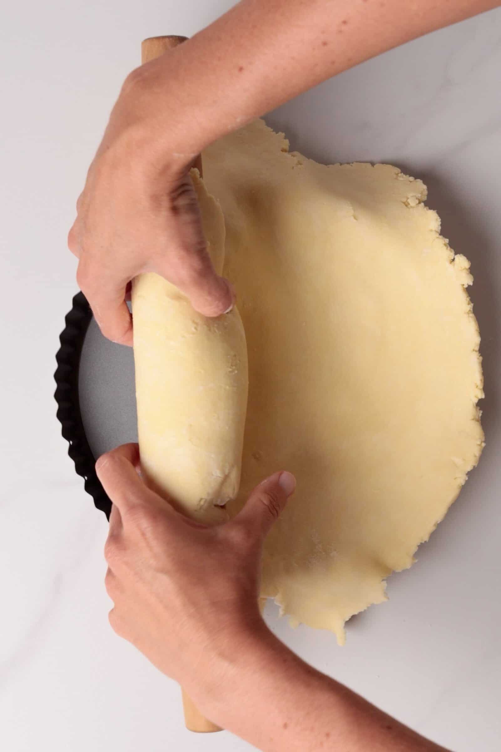 Hands unrolling pie dough from a rolling pin over a round tart pan, preparing to fit the dough into the pan on a white surface.