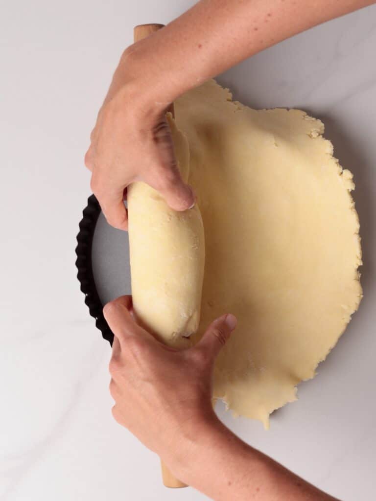 A person’s hands use a rolling pin to lift and place rolled-out pie dough into a tart pan on a light-colored surface.