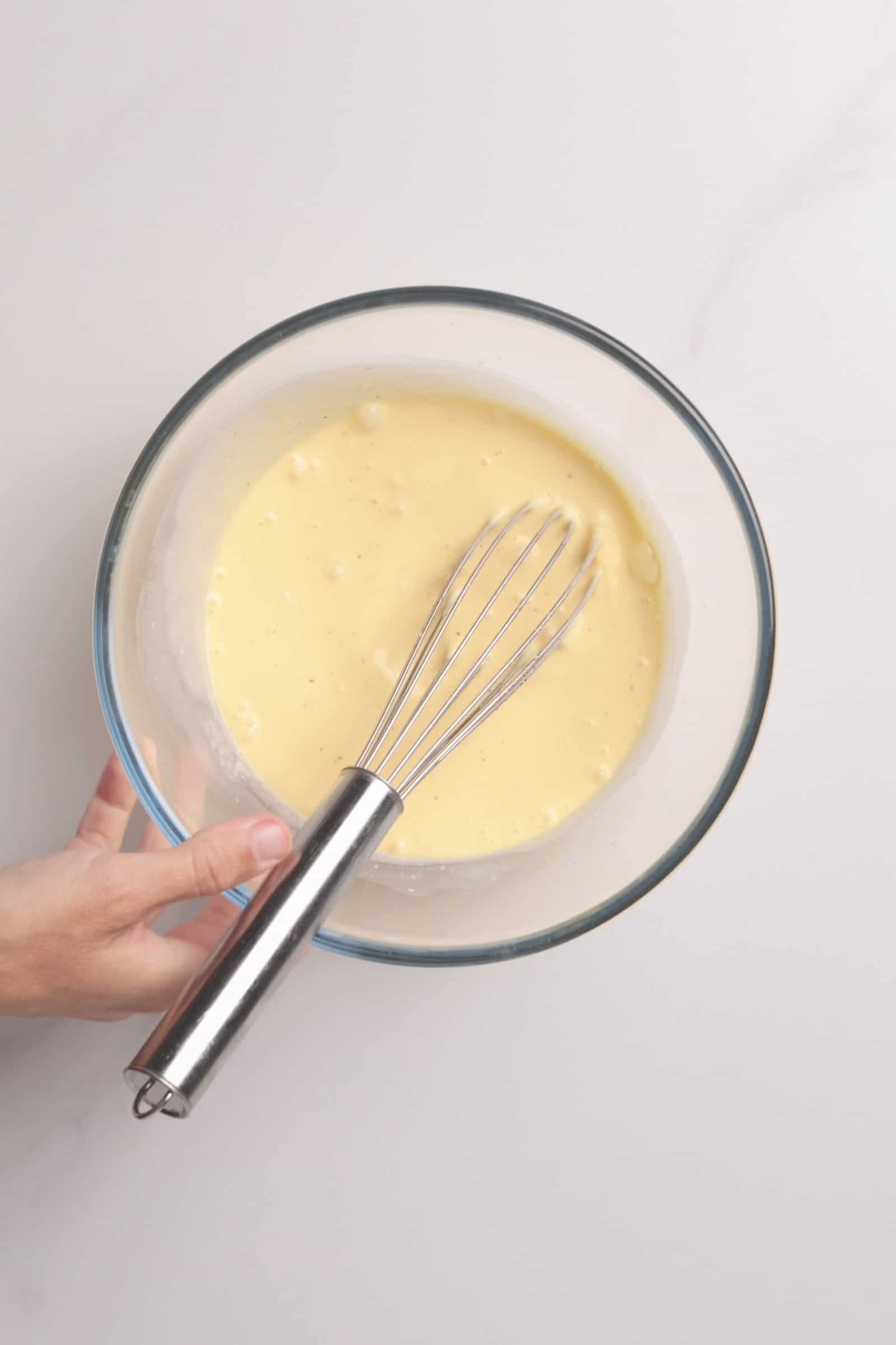 A hand holding a glass bowl filled with a light yellow batter, with a metal whisk resting inside, on a white surface.
