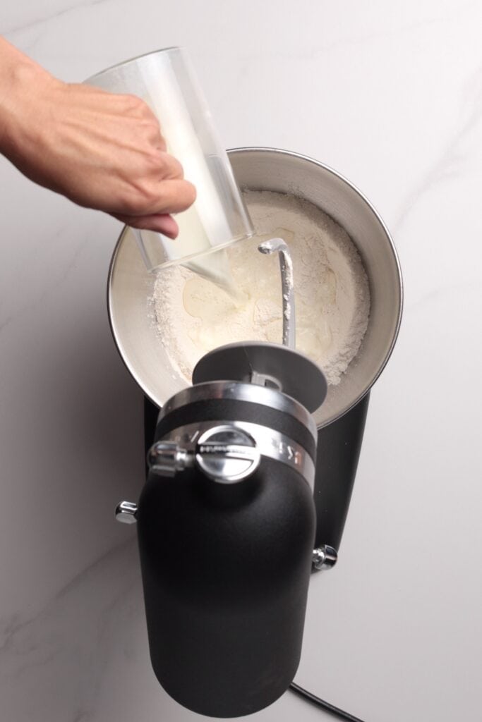 A hand pours milk from a clear measuring cup into a stand mixer bowl containing flour; the mixer has a dough hook attachment and is set on a light marble countertop.