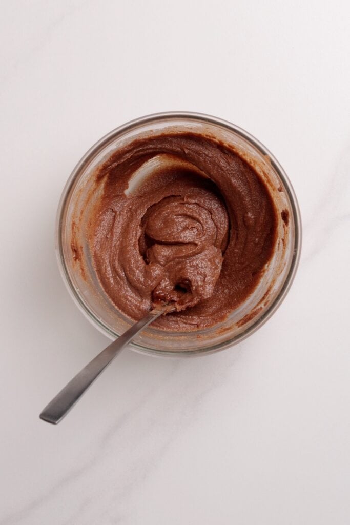 A glass bowl with creamy cinnamon batter being mixed, with a metal spoon resting inside, on a white marble surface.