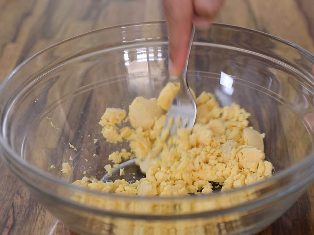 A hand mashes hard-boiled egg yolks with a fork in a clear glass bowl on a wooden surface.