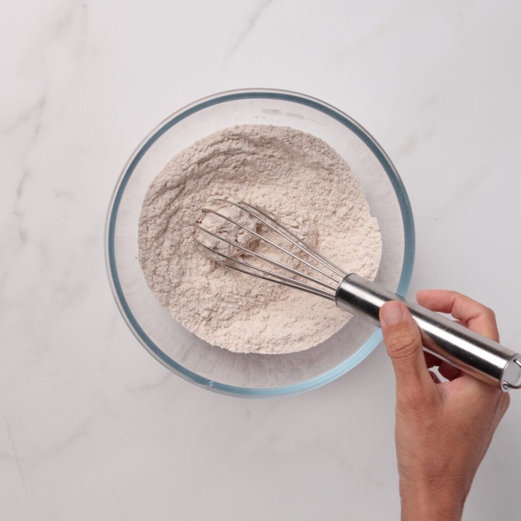 A hand holds a metal whisk, stirring flour in a clear glass bowl on a white marble surface.
