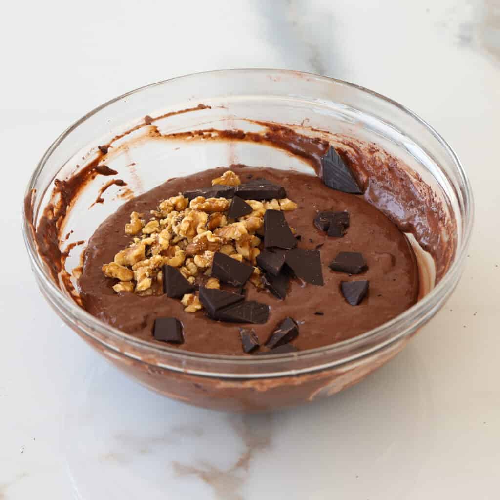 A glass bowl filled with chocolate cake batter, topped with chopped walnuts and pieces of dark chocolate, sits on a white marble surface.