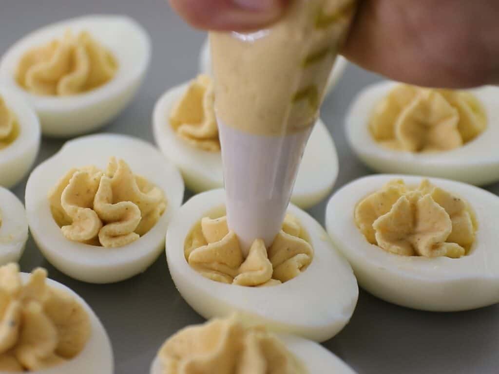 A hand uses a piping bag to fill halved hard-boiled eggs with a creamy, yellow deviled egg mixture. Several finished deviled eggs are visible on a gray surface.