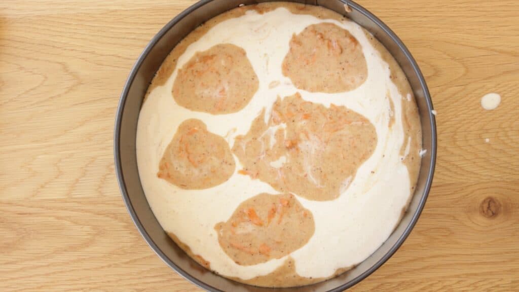 A round baking pan on a wooden surface, filled with a creamy white batter and five large dollops of light brown batter swirled on top, ready to be mixed or baked.