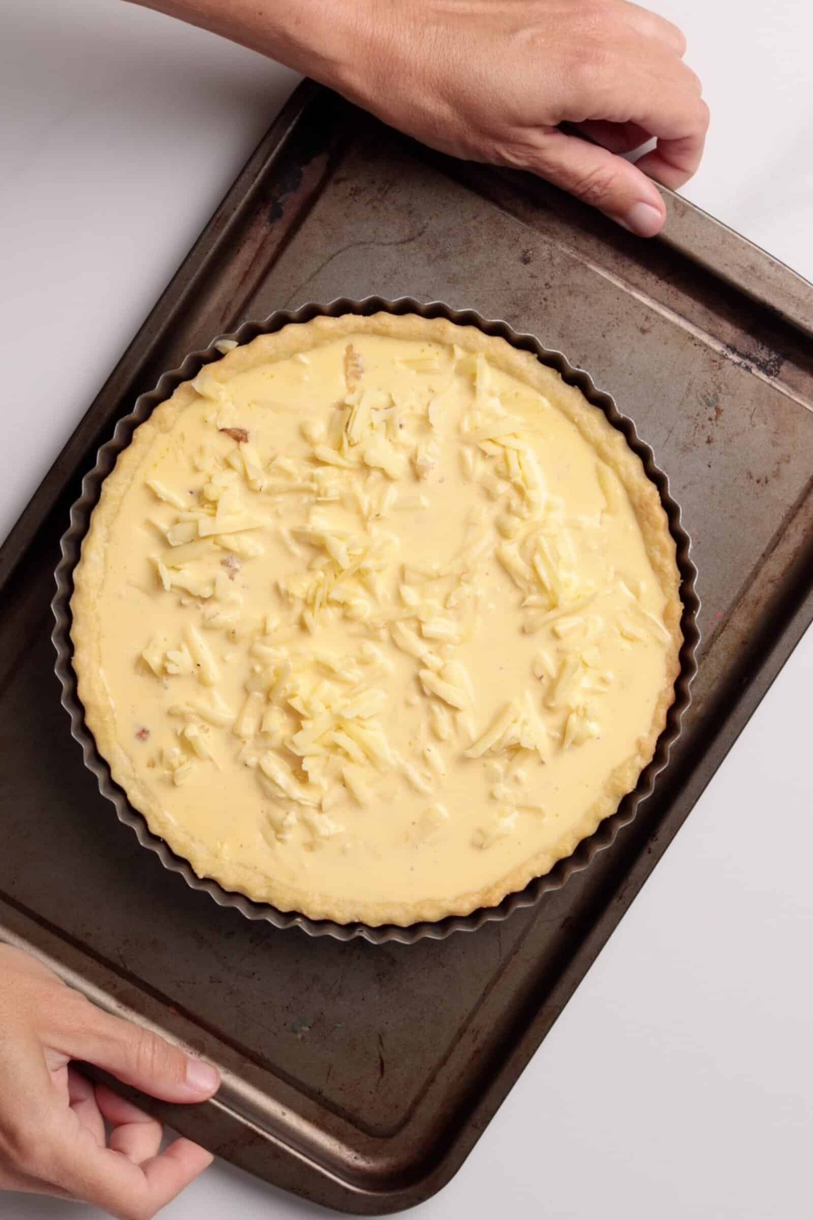 A person holds a baking tray with an unbaked quiche topped with shredded cheese, ready to go into the oven.