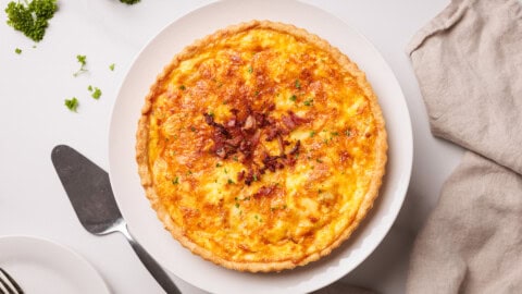 A freshly baked quiche with a golden, flaky crust and a cheesy, slightly browned filling, garnished with herbs and bacon bits, sits on a white plate next to a serving spatula and a beige napkin.