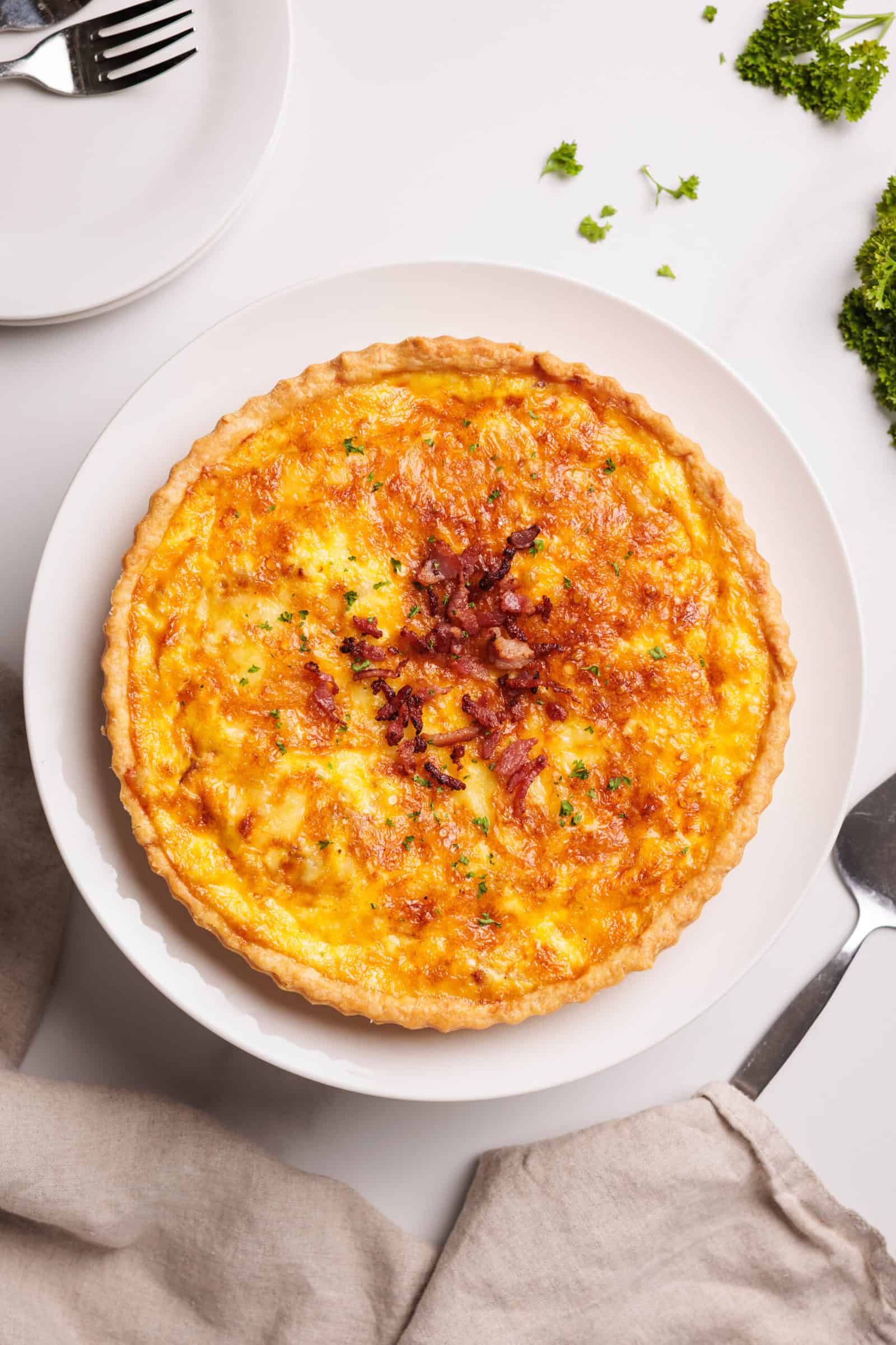 A golden-brown quiche topped with bits of bacon sits on a white plate, garnished with chopped herbs. A fork, knife, and napkin are nearby, with parsley leaves and empty plates visible in the background.