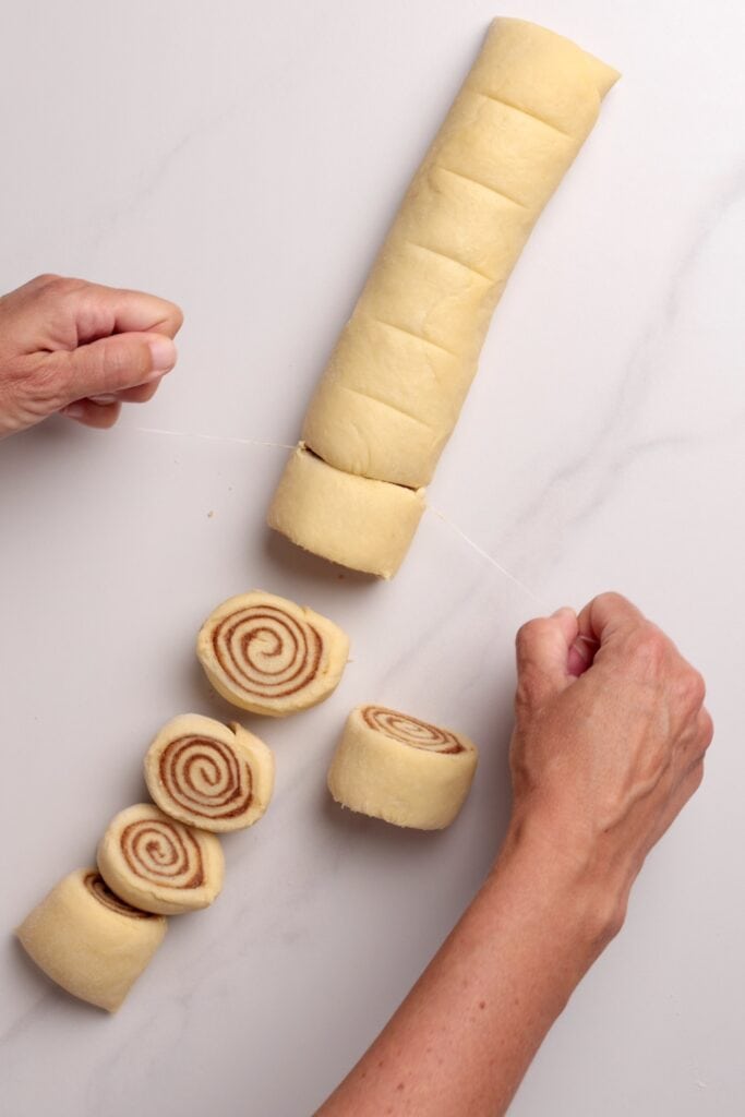 Hands use a piece of string to slice a rolled log of cinnamon roll dough into individual swirled pieces on a light surface.