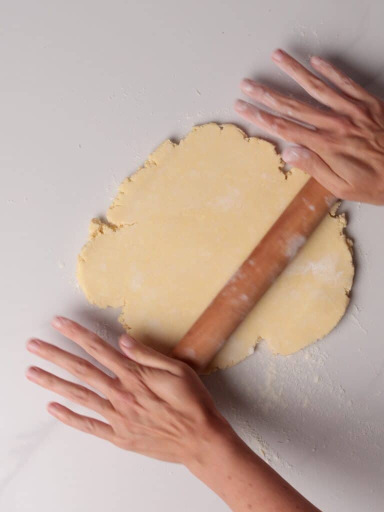 Two hands rolling out dough with a wooden rolling pin on a lightly floured surface.