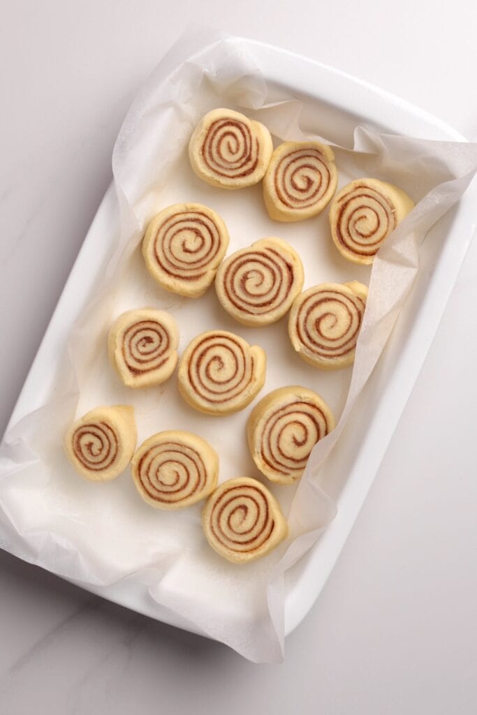 A white baking dish lined with parchment paper holds 12 unbaked cinnamon rolls arranged neatly in rows, ready to be baked.