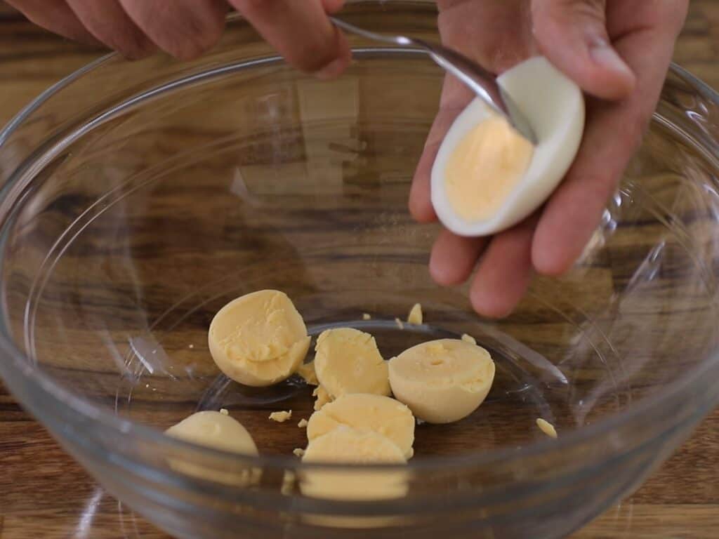 A person uses a spoon to scoop the yolk from a halved hard-boiled egg over a glass bowl containing other egg halves and yolks.