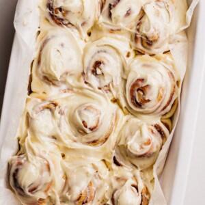 A tray of freshly baked cinnamon rolls covered in creamy white icing, arranged closely together on parchment paper inside a white baking dish.