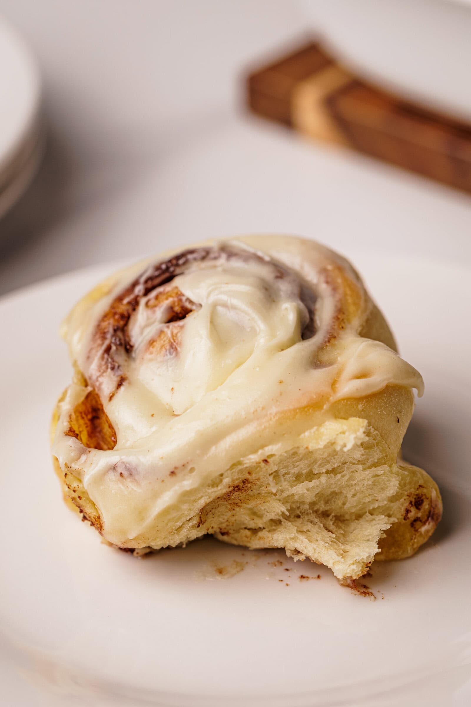 A close-up of a cinnamon roll topped with creamy icing sits on a white plate, with a bite taken out of it, revealing the soft, fluffy texture inside.