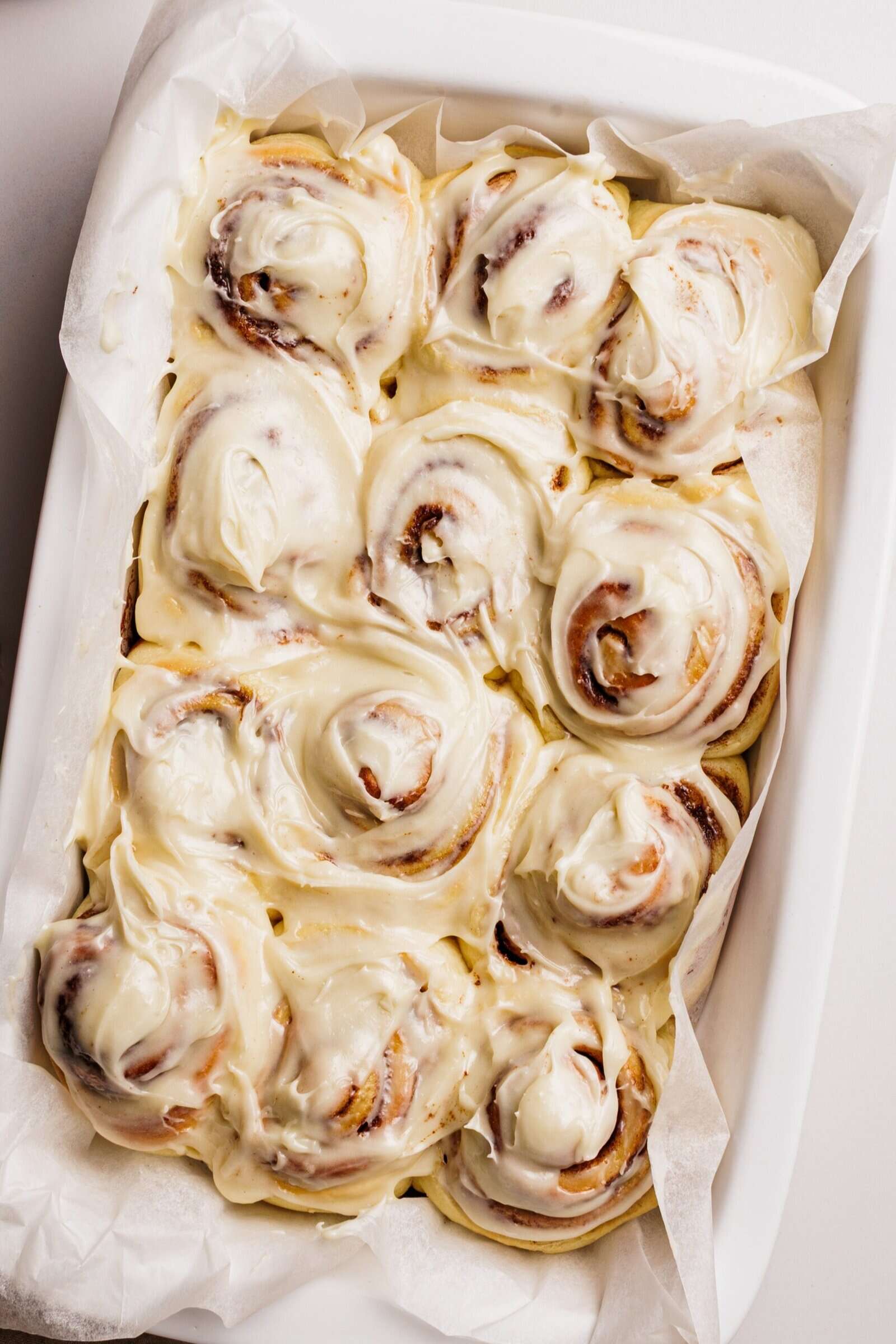 A baking dish lined with parchment paper holds nine frosted cinnamon rolls, swirled and generously covered in creamy cream cheese frosting.