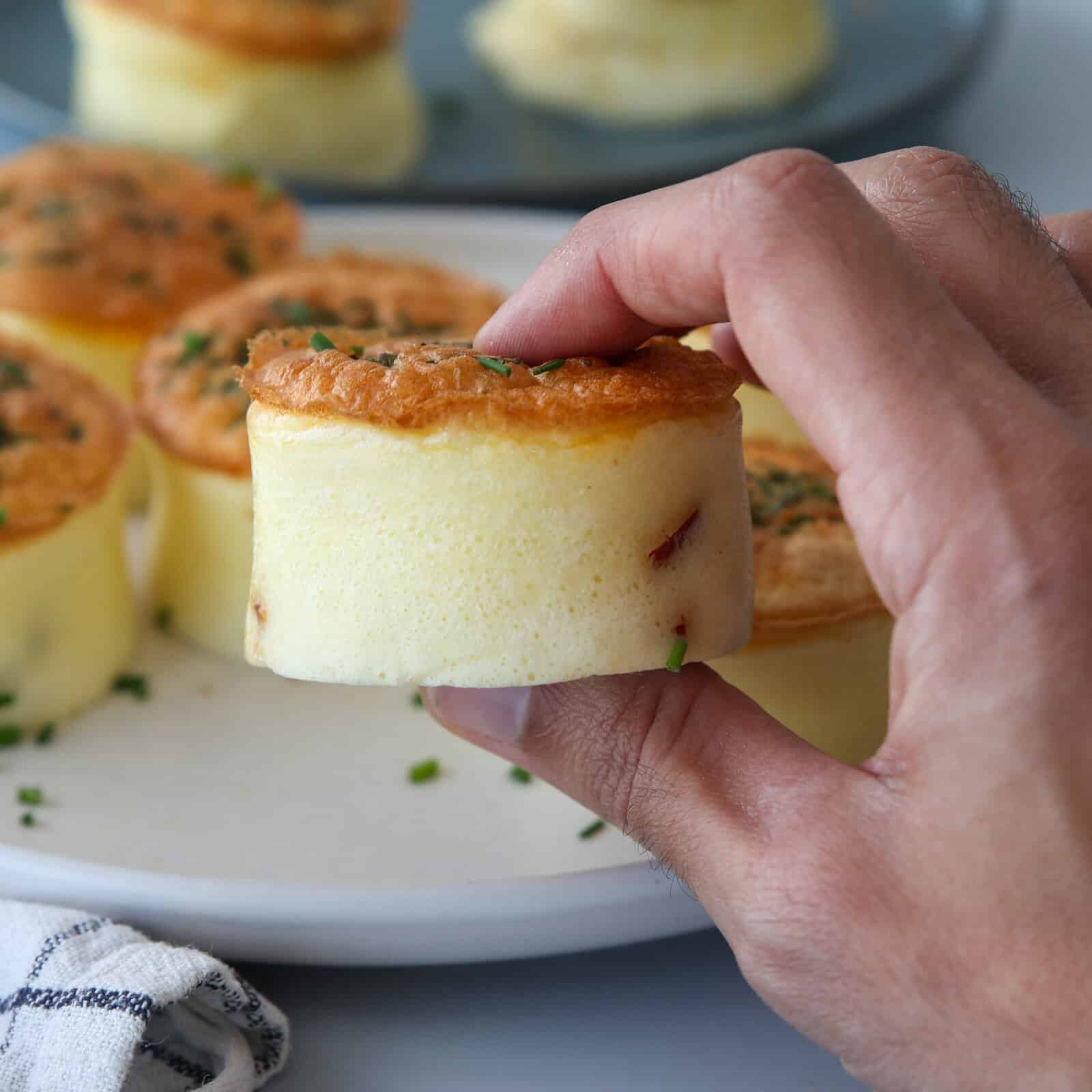 A hand holding a fluffy, round egg bite garnished with chopped herbs, with more soufflés on a white plate in the background.