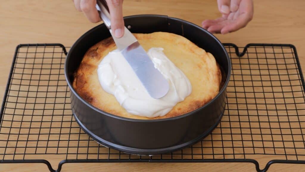 A person spreads a layer of white cream on a round cheesecake in a springform pan, placed on a cooling rack on a wooden surface.
