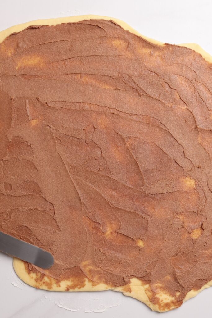 A rolled-out sheet of dough is being spread with a layer of cinnamon sugar filling using a spatula on a white surface.