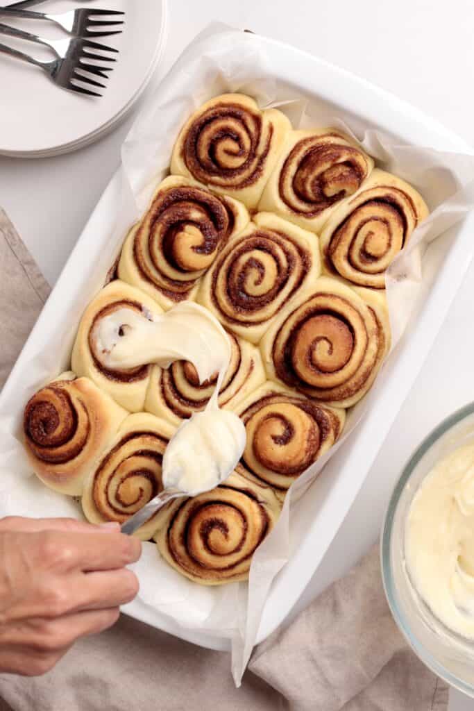 A hand spreads creamy icing onto freshly baked cinnamon rolls in a white baking dish. A bowl of icing, stacked plates, and forks are nearby on a light surface.