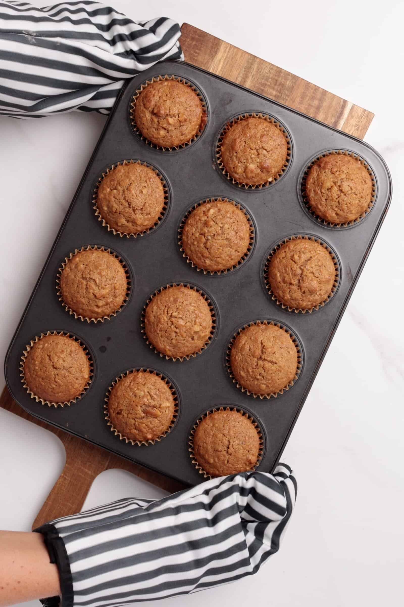 A person wearing black and white striped oven mitts holds a muffin tin filled with twelve freshly baked muffins on a wooden cutting board.
