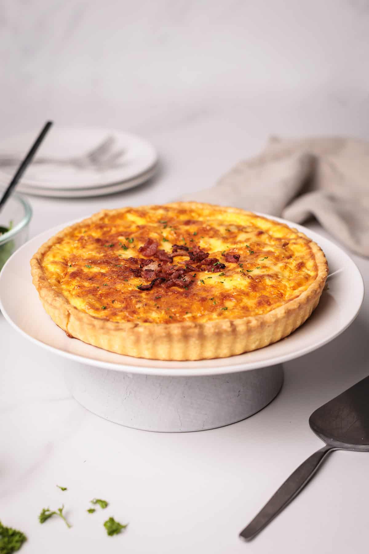 A baked quiche Lorraine with a golden, flaky crust and a savory filling topped with bacon bits sits on a white cake stand. Plates, cutlery, a napkin, and sprigs of parsley are in the background.