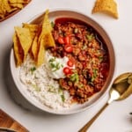 A bowl with white rice, chili topped with sliced red chili peppers, chopped cilantro, and sour cream, served with tortilla chips on the side. A gold spoon and extra tortilla chips are nearby on a white surface.