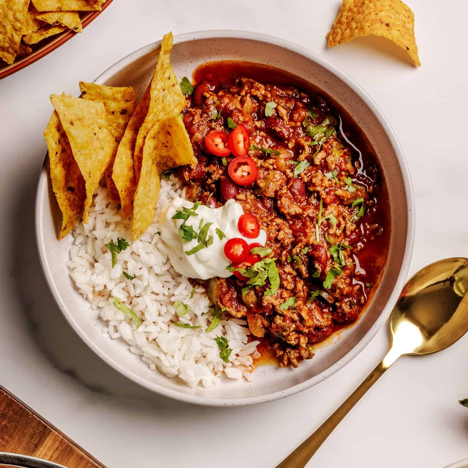 A bowl with white rice, chili topped with sliced red chili peppers, chopped cilantro, and sour cream, served with tortilla chips on the side. A gold spoon and extra tortilla chips are nearby on a white surface.